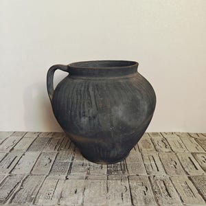 May include: A large, dark gray ceramic jug with a single handle. The jug has a rounded body and a wide opening. The surface has a textured, weathered appearance, suggesting age. The jug is likely for decorative or functional use.