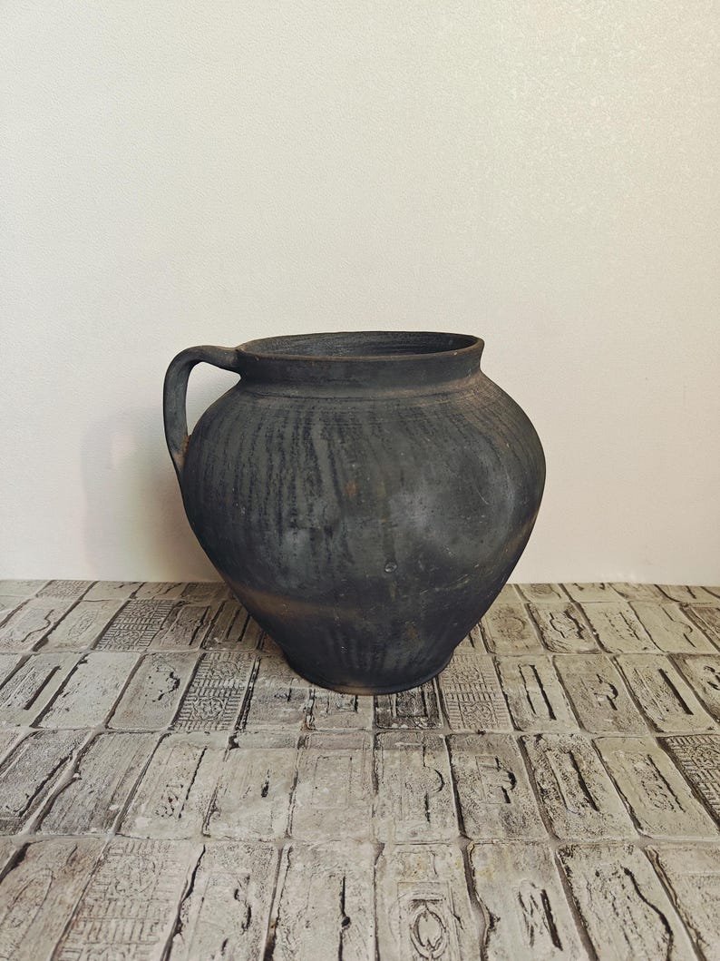 May include: A large, dark gray ceramic jug with a single handle. The jug has a rounded body and a wide opening. The surface has a textured, weathered appearance, suggesting age. The jug is likely for decorative or functional use.