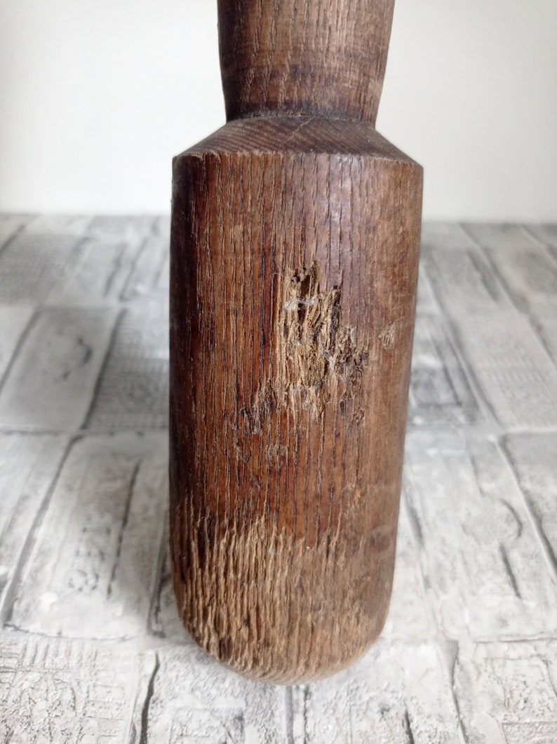 May include: A close-up of a wooden rolling pin with a worn, rustic finish. The pin has a round shape and is made of dark brown wood with visible grain patterns. There are signs of wear and tear on the surface, including a small knot and a few scratches.