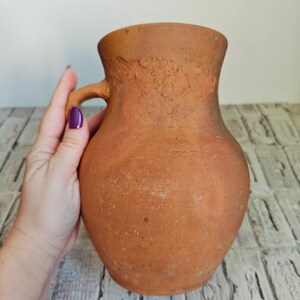 May include: A rustic, terracotta-colored pitcher with a handle. The pitcher has a rounded body and a slightly flared rim. The surface has a textured, aged appearance, suggesting it is handmade. The handle is small and curved, and the overall design is simple and functional.