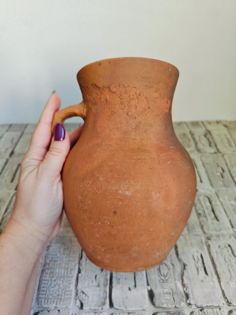 May include: A rustic, terracotta-colored pitcher with a handle. The pitcher has a rounded body and a slightly flared rim. The surface has a textured, aged appearance, suggesting it is handmade. The handle is small and curved, and the overall design is simple and functional.