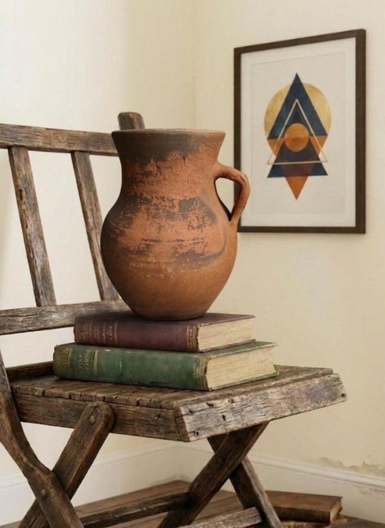 May include: A rustic, weathered wooden chair holds a stack of vintage books and a terracotta vase. The vase has a handle and sits atop the books. A framed geometric art piece hangs in the background.