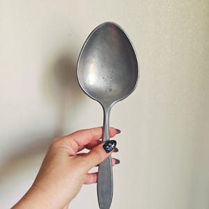 May include: A large, vintage-style metal spoon with a wide, oval bowl and a long, flat handle. The spoon has a matte, silver finish and shows signs of wear. The spoon is held against a neutral background.