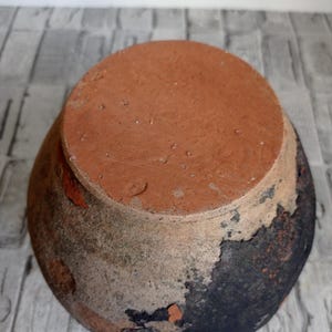 May include: A terracotta pot with a dark brown glaze. The pot has a round shape and a wide opening. The glaze is chipped and cracked in places, revealing the terracotta underneath.