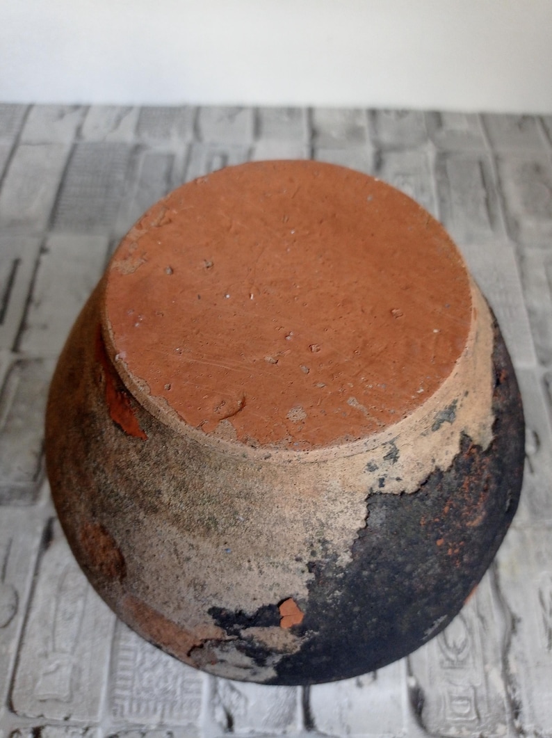 May include: A terracotta pot with a dark brown glaze. The pot has a round shape and a wide opening. The glaze is chipped and cracked in places, revealing the terracotta underneath.