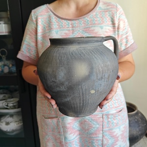 Op de afbeelding: Een witte keramische mok met een zwarte handgreep en de tekst "But First, Coffee" in een vetgedrukte, zwarte letter. De mok staat tegen een vage achtergrond, wat een gezellige setting suggereert.