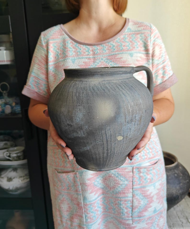 May include: A large, dark gray ceramic jug with a handle. The jug has a rounded body and a textured surface. The background includes a patterned dress and a glimpse of other pottery. This rustic piece is suitable for home decor.