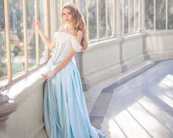 May include: A woman wearing a light blue gown with a white lace bodice and a tiara. She is standing in a glass building with a white background.