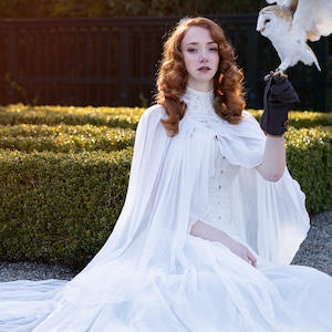 May include: A woman wearing a white gown and cape sits in a garden with a white owl perched on her gloved hand.