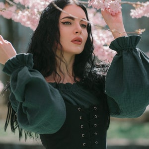 May include: A woman with long black hair is wearing a dark green blouse with puffed sleeves and a black lace-up corset. She is standing in front of a pink cherry blossom tree.