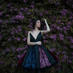 May include: A woman in a black, blue, and purple dress with a geometric pattern stands in front of a wall of purple flowers.