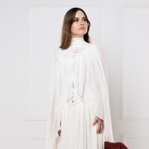 May include: A white, long-sleeved dress with a high neck and a flowing skirt. The dress has a lace overlay and a belt at the waist. The model is standing in front of a white wall.