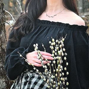 May include: A woman with long dark hair wearing a black off-the-shoulder top with a ruffled neckline and a black and white plaid skirt. She is holding a bouquet of white pussy willow branches.