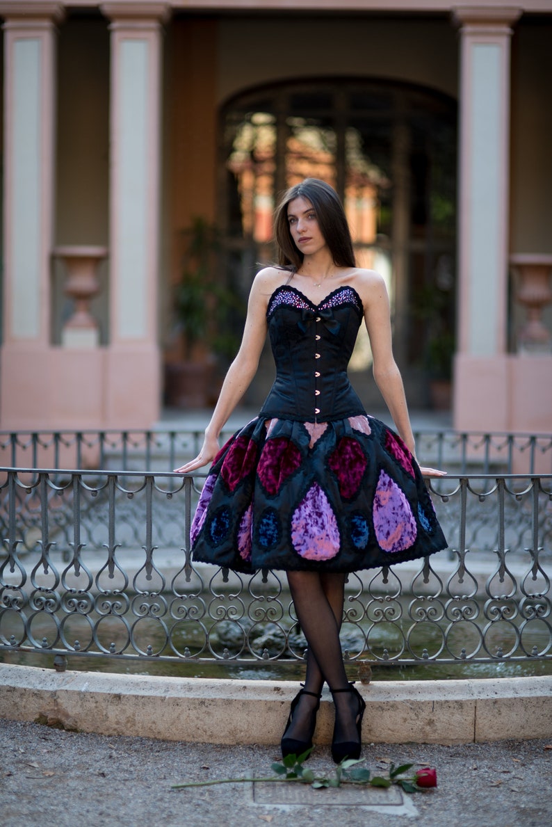 K&ouml;nnte beinhalten: Eine Frau tr&auml;gt ein schwarzes Korsett mit einem rosa-violetten gemusterten Rock. Sie steht vor einem schmiedeeisernen Zaun.