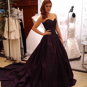 May include: A floor-length strapless gown in a deep dark purple hue featuring a fitted sweetheart bodice and a voluminous, pleated skirt that extends into a long train. The garment is constructed with structured seams along the torso.