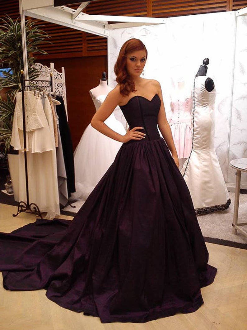May include: A floor-length strapless gown in a deep dark purple hue featuring a fitted sweetheart bodice and a voluminous, pleated skirt that extends into a long train. The garment is constructed with structured seams along the torso.