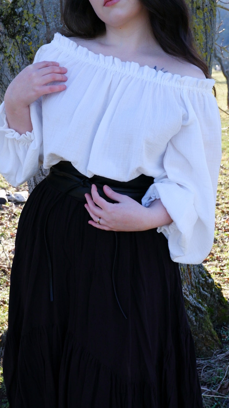 May include: A white off-the-shoulder blouse with long sleeves and a black leather belt. The blouse is gathered at the neckline and the sleeves are puffed. The belt is tied at the waist.