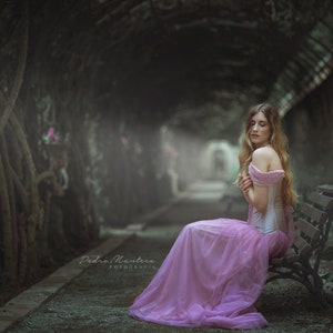 May include: A woman in a pink dress sits on a wooden bench in a garden setting. The bench is surrounded by lush greenery and the woman is looking down with a thoughtful expression.