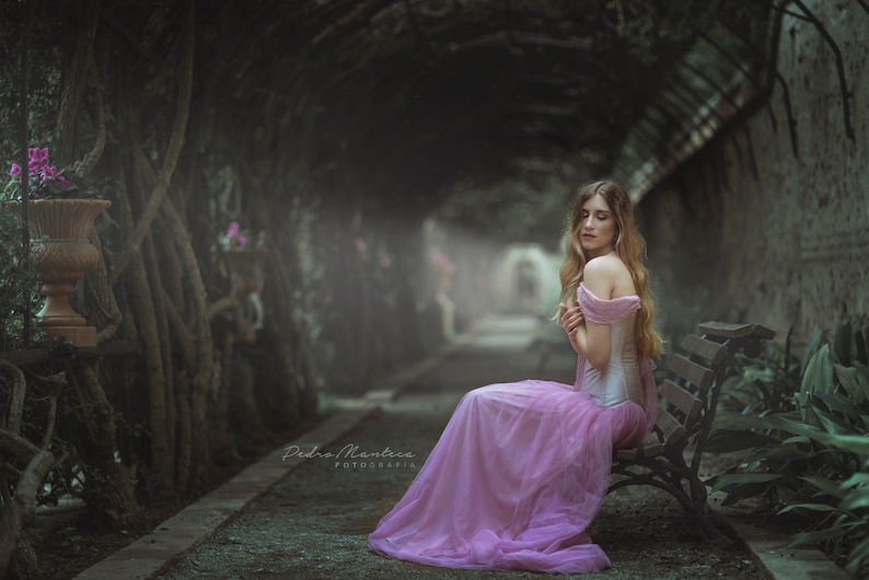 May include: A woman in a pink dress sits on a wooden bench in a garden setting. The bench is surrounded by lush greenery and the woman is looking down with a thoughtful expression.