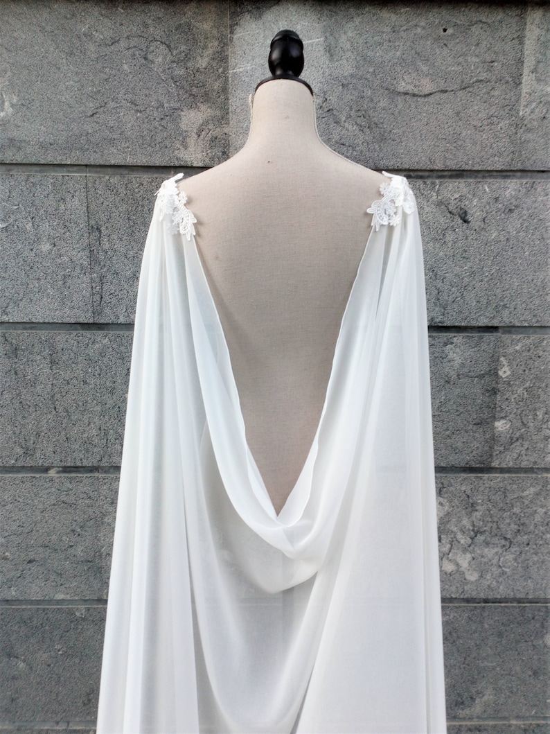 Pu&ograve; includere: Cappotto da sposa in chiffon bianco con dettagli in pizzo sulle spalle. Il mantello &egrave; drappeggiato su un manichino e ha una schiena bassa.