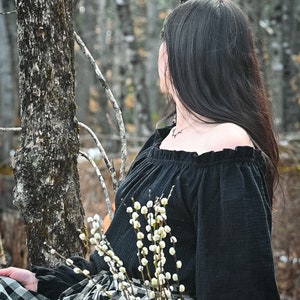 May include: A woman wearing a black off-the-shoulder top with a ruffled neckline and a black and white checkered skirt. She is holding a bouquet of white pussy willow branches.