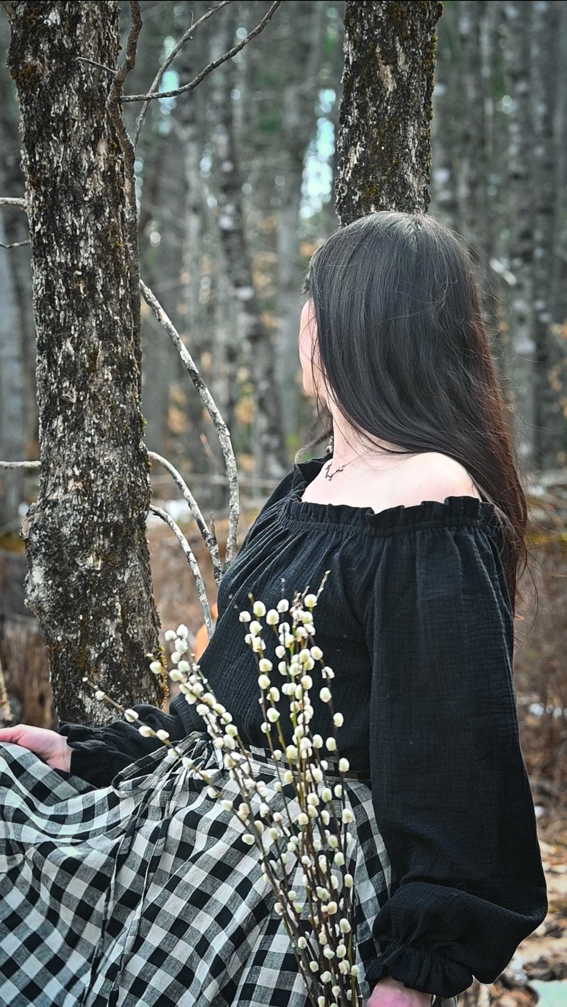 May include: A woman wearing a black off-the-shoulder top with a ruffled neckline and a black and white checkered skirt. She is holding a bouquet of white pussy willow branches.