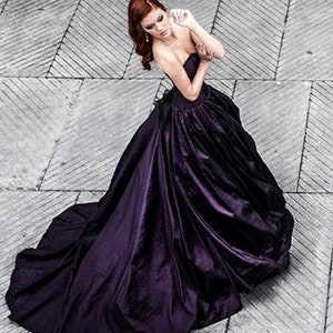 May include: A floor-length strapless gown in a deep, dark purple hue. The dress features a fitted bodice and a voluminous, full skirt with a long train that pools on the ground. The fabric has a subtle sheen, creating soft folds and shadows.