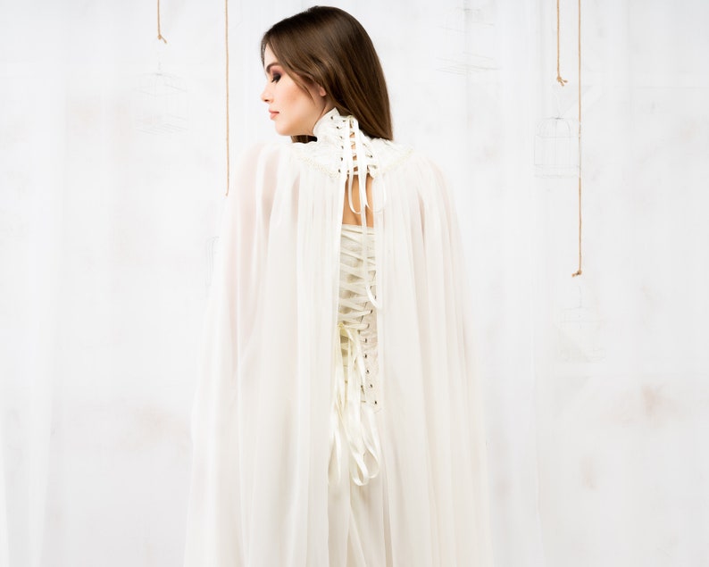May include: A white sheer cape with a white lace-up corset. The cape is flowing and the corset is cinched at the waist.