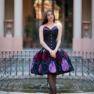 K&ouml;nnte beinhalten: Eine Frau tr&auml;gt ein schwarzes Korsett mit einem Rock mit violettem und blauem Muster. Sie steht vor einem schmiedeeisernen Zaun.