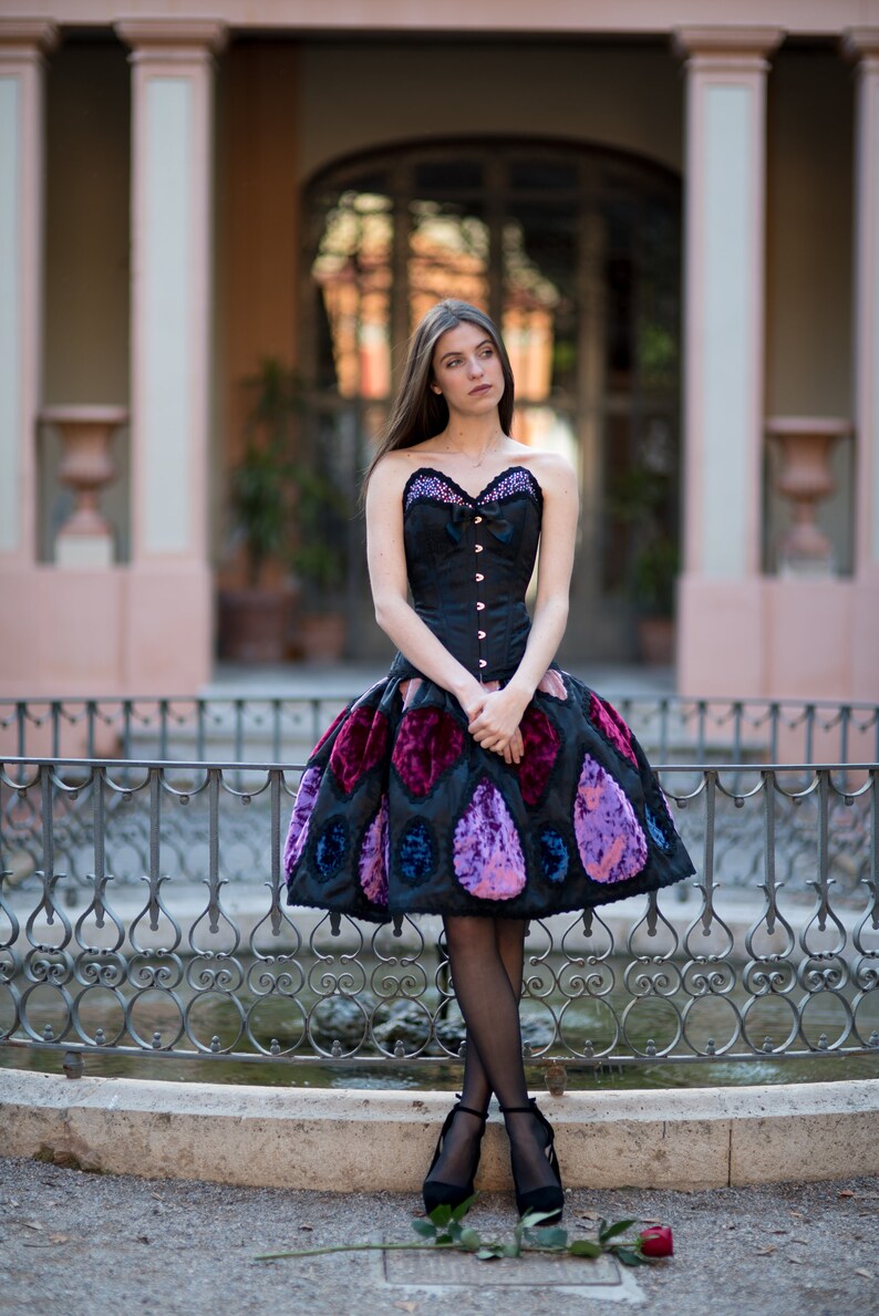 K&ouml;nnte beinhalten: Eine Frau tr&auml;gt ein schwarzes Korsett mit einem Rock mit violettem und blauem Muster. Sie steht vor einem schmiedeeisernen Zaun.