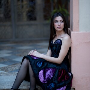 K&ouml;nnte beinhalten: Eine Frau in einem schwarzen Samtkleid mit violetten und blauen Paillettenbesatz. Sie sitzt auf einer Treppe mit einer pinkfarbenen Wand hinter ihr.