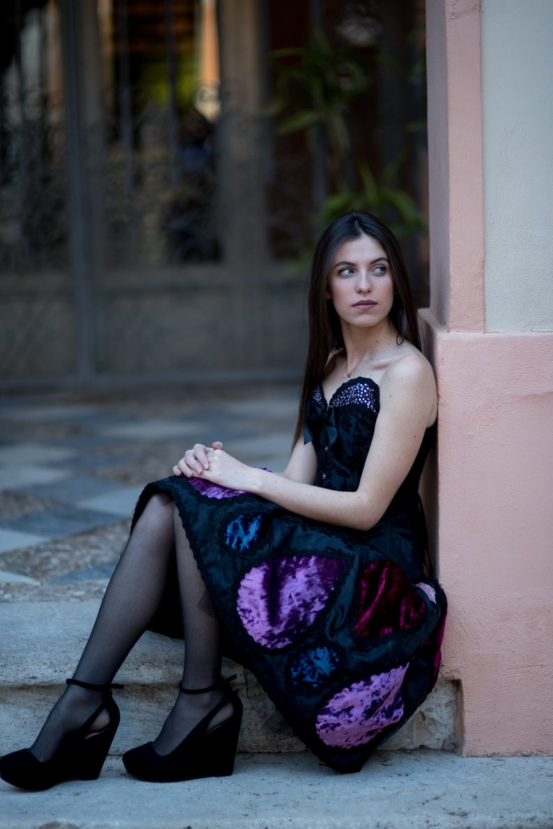 K&ouml;nnte beinhalten: Eine Frau in einem schwarzen Samtkleid mit violetten und blauen Paillettenbesatz. Sie sitzt auf einer Treppe mit einer pinkfarbenen Wand hinter ihr.