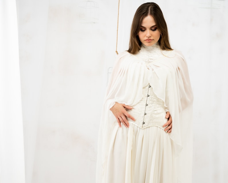 May include: A woman wearing a white, flowing dress with a lace corset. The dress has a high neckline and long sleeves. The woman is standing in front of a white background.