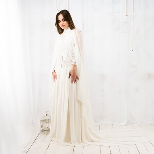 May include: A woman wearing a white gown with a long train. The gown has a high neckline and a fitted bodice. The woman is standing in a white room with a white background.
