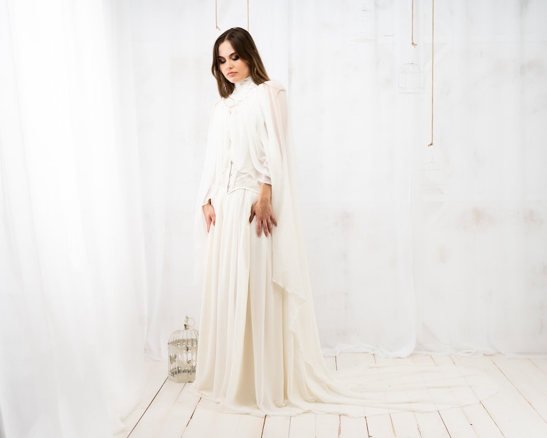 May include: A woman wearing a white gown with a long train. The gown has a high neckline and a fitted bodice. The woman is standing in a white room with a white background.