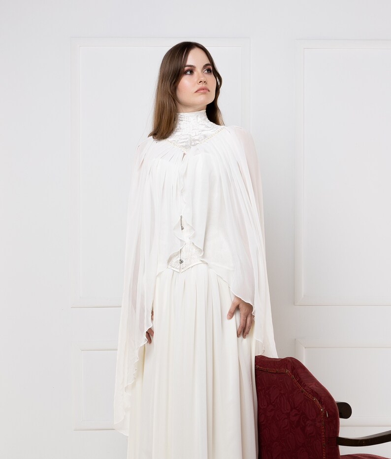 May include: A white, long-sleeved dress with a high neck and a flowing skirt. The dress has a lace overlay and a belt at the waist. The model is standing in front of a white wall.