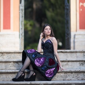 K&ouml;nnte beinhalten: Eine Frau in einem schwarzen Kleid mit violetten und blauen Samtdetails sitzt auf einer Steintreppe. Das Kleid hat einen herzf&ouml;rmigen Ausschnitt und ein tailliertes Oberteil. Sie tr&auml;gt schwarze Abs&auml;tze und hat ihre Haare nach hinten gebunden.