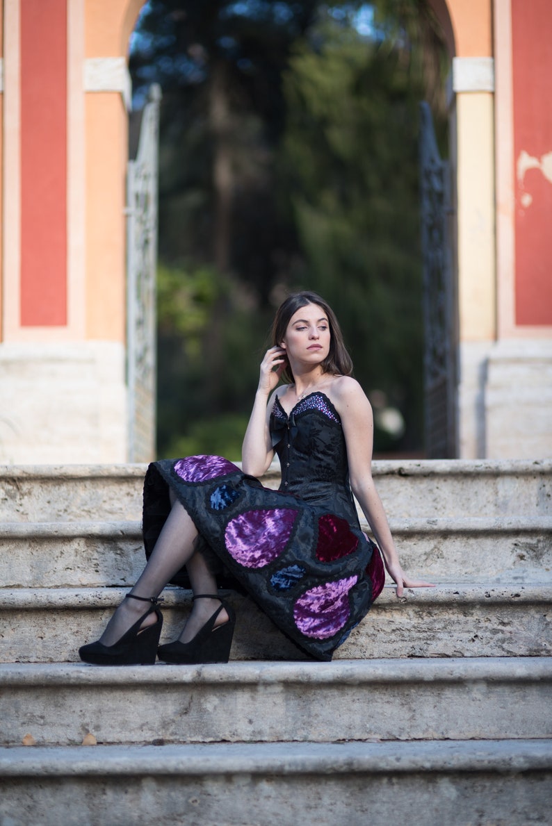 K&ouml;nnte beinhalten: Eine Frau in einem schwarzen Kleid mit violetten und blauen Samtdetails sitzt auf einer Steintreppe. Das Kleid hat einen herzf&ouml;rmigen Ausschnitt und ein tailliertes Oberteil. Sie tr&auml;gt schwarze Abs&auml;tze und hat ihre Haare nach hinten gebunden.