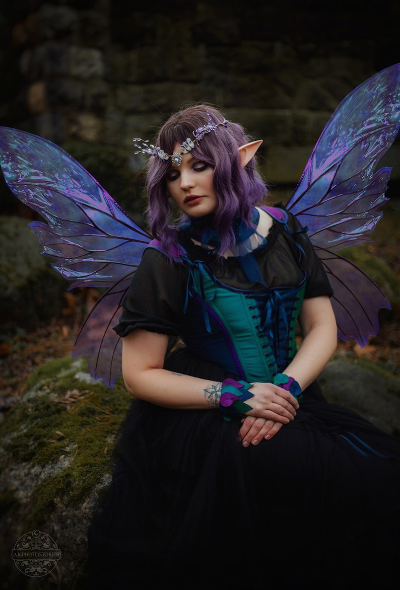 May include: A woman with purple hair and iridescent fairy wings sits on a mossy rock. She is wearing a black dress, a teal corset, and a silver crown with purple flowers.