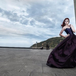 May include: A floor-length, strapless gown in a deep shade of dark purple. The bodice is fitted and structured, transitioning into a voluminous, full-length skirt with subtle folds and a smooth, slightly lustrous fabric texture throughout the garment.