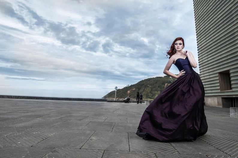 May include: A floor-length, strapless gown in a deep shade of dark purple. The bodice is fitted and structured, transitioning into a voluminous, full-length skirt with subtle folds and a smooth, slightly lustrous fabric texture throughout the garment.