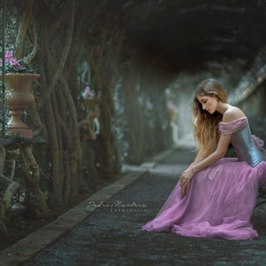 May include: A young woman in a pink dress sits on a bench in a tunnel of trees. The trees are overgrown and the tunnel is dark and mysterious. The woman is looking down and her hair is long and flowing.
