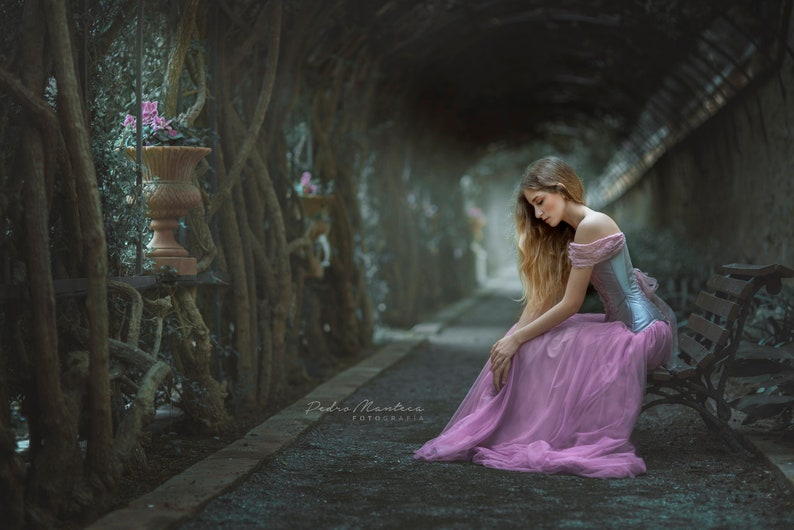 May include: A young woman in a pink dress sits on a bench in a tunnel of trees. The trees are overgrown and the tunnel is dark and mysterious. The woman is looking down and her hair is long and flowing.