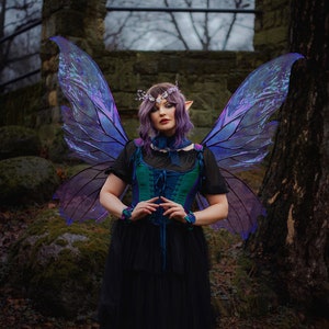 May include: A woman wearing a dark green and blue corset with a black tulle skirt and iridescent purple butterfly wings. She is standing in a forest setting with a stone wall and trees in the background.