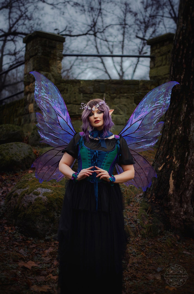 May include: A woman wearing a dark green and blue corset with a black tulle skirt and iridescent purple butterfly wings. She is standing in a forest setting with a stone wall and trees in the background.
