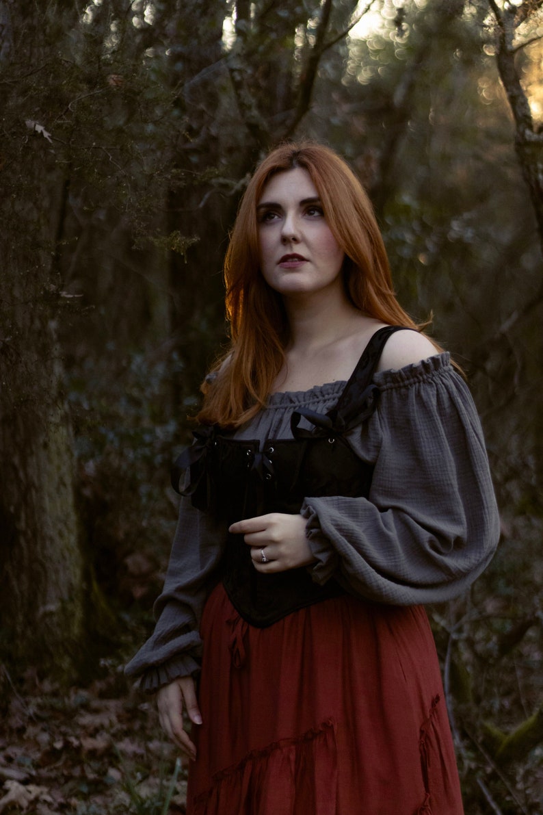 May include: A woman with long red hair wearing a gray off-the-shoulder blouse with a black lace-up corset and a red skirt with ruffles.