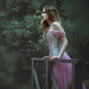 May include: A woman in a pink dress stands on a wooden bridge in a forest. She is looking off to the side and her hair is long and flowing. The background is blurred and the image has a dreamy, ethereal feel.
