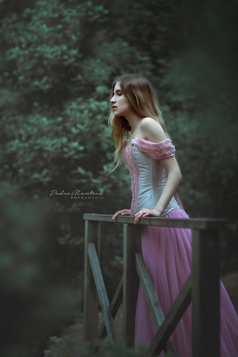 May include: A woman in a pink dress stands on a wooden bridge in a forest. She is looking off to the side and her hair is long and flowing. The background is blurred and the image has a dreamy, ethereal feel.