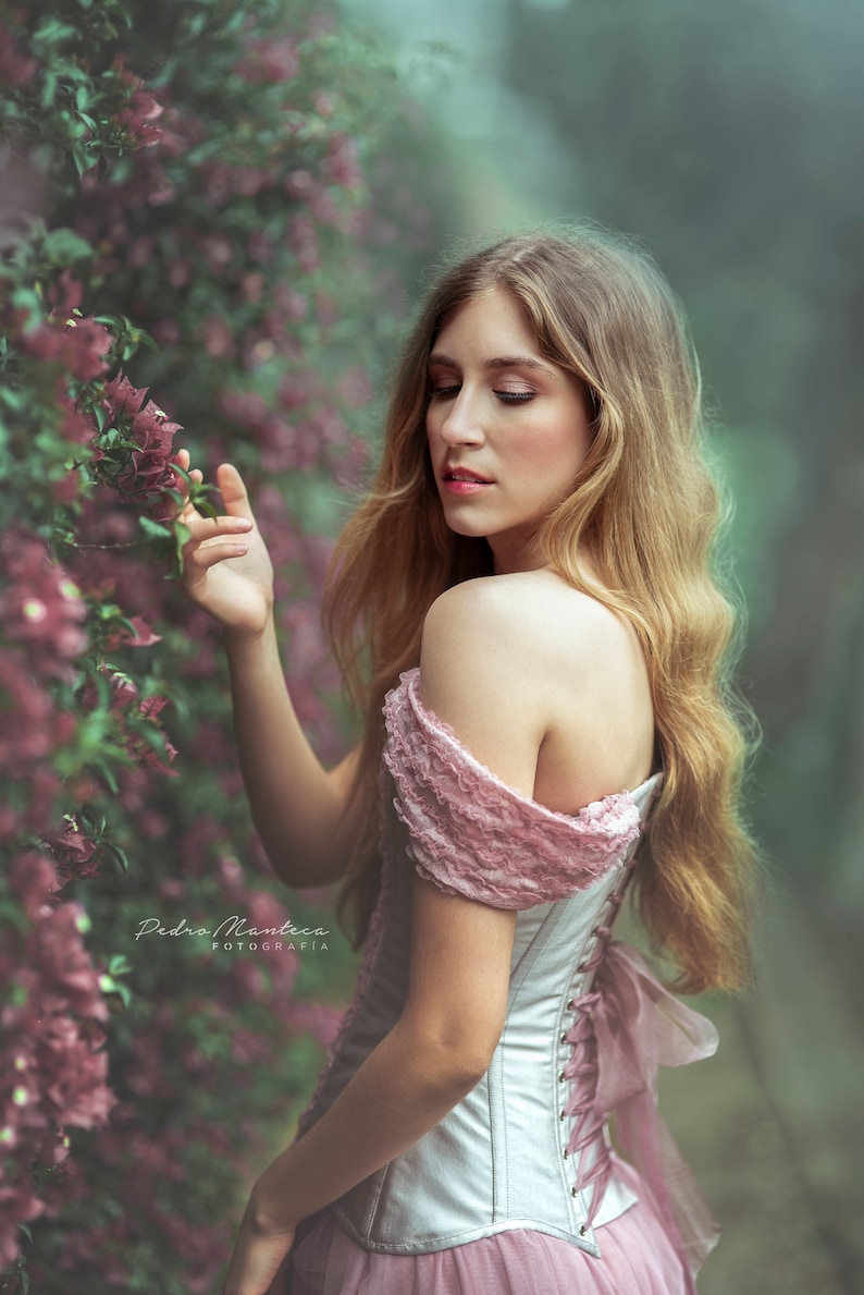 May include: A woman with long blonde hair wears a white corset with a pink lace overlay and a pink skirt. She is standing in front of a wall of pink flowers.