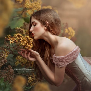 May include: A woman in a pink and white dress with ruffles is smelling yellow flowers. The flowers are in focus and the woman is out of focus. The background is blurred and has a warm, golden tone.